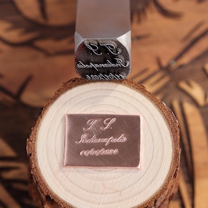 Könnte beinhalten: Ein Metallstempel mit den Initialen "R.R." und anderem Text befindet sich über einer Kupferplatte, die mit "T.S. Indianapolis 11/01/2020" graviert ist. Die Platte liegt auf einer Holzscheibe und zeigt einen personalisierten Gravurprozess.