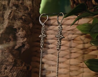 May include: Silver-toned dangle earrings with a cross-shaped design and chain detail. The earrings have a hook closure and a small, round charm at the end of the chain. The background includes a woven texture and greenery.