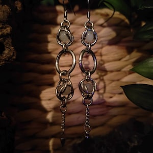 Puede incluir: Un par de pendientes largos plateados. Cada pendiente presenta una serie de anillos ovalados enlazados, pequeños detalles de cadena y una piedra redonda pálida encerrada en una jaula de alambre. Los pendientes cuelgan de un gancho simple.