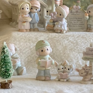 May include: A miniature Christmas village scene with a schoolhouse, a clock tower, a Christmas tree, and several figurines. The figurines are dressed in winter clothing and are holding gifts and other holiday items.