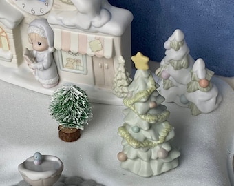 May include: A ceramic Christmas village scene with a church, street signs, and miniature trees. A dog figurine wearing a Santa hat sits on a bench next to a green wreath. The scene is set on a snowy landscape.