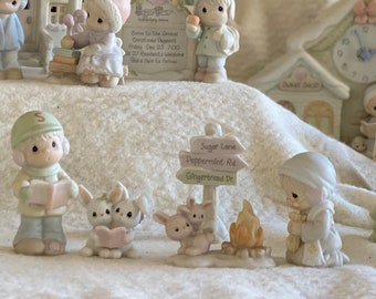 May include: A miniature Christmas village scene with a schoolhouse, a church, a Christmas tree, and several figurines. The figurines are dressed in winter clothing and are holding various items, such as presents, candy canes, and a book. The scene is set on a white, fluffy surface that resembles snow.