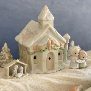 May include: A white porcelain Christmas village scene with a church, a manger, a Christmas tree, and two figures singing carols. The church has a heart-shaped window and a cross on the door. The manger has a star on top. The Christmas tree is decorated with ornaments. The figures are wearing winter clothes and holding a book.
