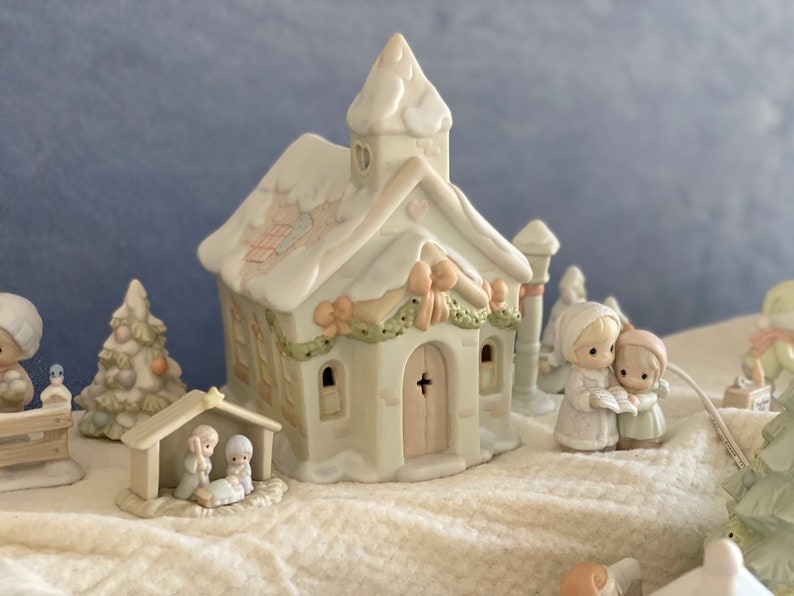 May include: A white porcelain Christmas village scene with a church, a manger, a Christmas tree, and two figures singing carols. The church has a heart-shaped window and a cross on the door. The manger has a star on top. The Christmas tree is decorated with ornaments. The figures are wearing winter clothes and holding a book.