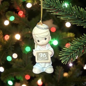 May include: A Christmas tree ornament featuring a cherubic angel holding a chalkboard with the number 96 and the words "Peace on Earth." The angel is wearing a white robe and blue pants, with a halo above its head. The ornament is hanging from a gold string.