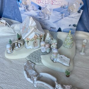 May include: A Precious Moments Sugar Town Christmas village scene with a church, a bridge, trees, and figurines. The sign reads "Welcome to Sugar Town".