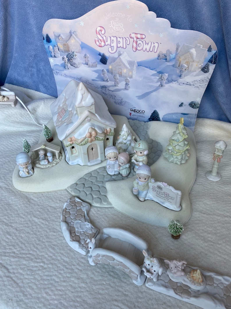 May include: A Precious Moments Sugar Town Christmas village scene with a church, a bridge, trees, and figurines. The sign reads "Welcome to Sugar Town".