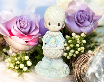 May include: A porcelain figurine of a boy holding a basket filled with pastel blue eggs and a small yellow chick. The figurine is surrounded by artificial flowers and greenery.