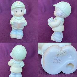 May include: A porcelain figurine of a child wearing a green hat and a blue coat, holding a book. The figurine is part of the Precious Moments collection and is marked "Philip &copy;1992 PMI 529494 Thailand".