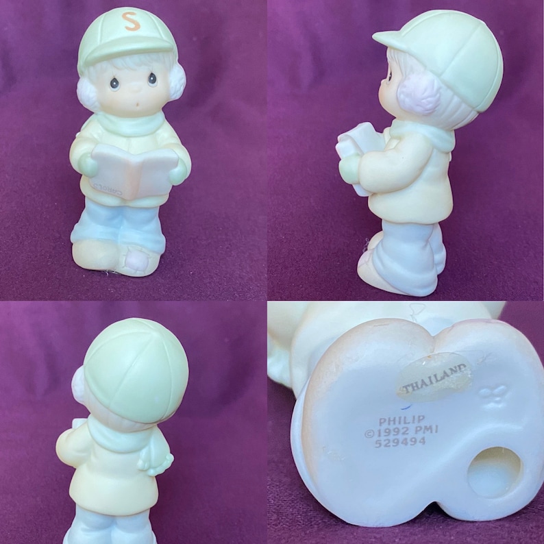 May include: A porcelain figurine of a child wearing a green hat and a blue coat, holding a book. The figurine is part of the Precious Moments collection and is marked "Philip &copy;1992 PMI 529494 Thailand".