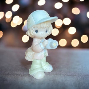 May include: A small figurine of a child wearing a white hat and holding a camera. The figurine is made of white porcelain and has a light pink and yellow color scheme. The background is blurred and features a bokeh effect of warm white lights.