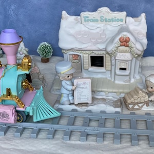 May include: A pastel pink and blue toy train engine with gold accents pulls a flatbed car on a gray track. The train is in front of a white snow-covered train station with a red bow and the words "Train Station" on the sign. A small figurine of a person in a blue uniform stands next to a sign that says "Passenger List". A small figurine of a person in a white coat is pushing a wooden sled with logs on it.