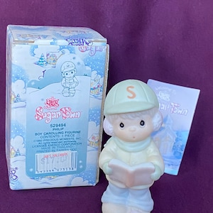 May include: A porcelain figurine of a boy wearing a green hat with the letter "S" on it, a blue jacket, and blue pants. He is holding a book and singing carols. The figurine is part of the Precious Moments Sugar Town collection.