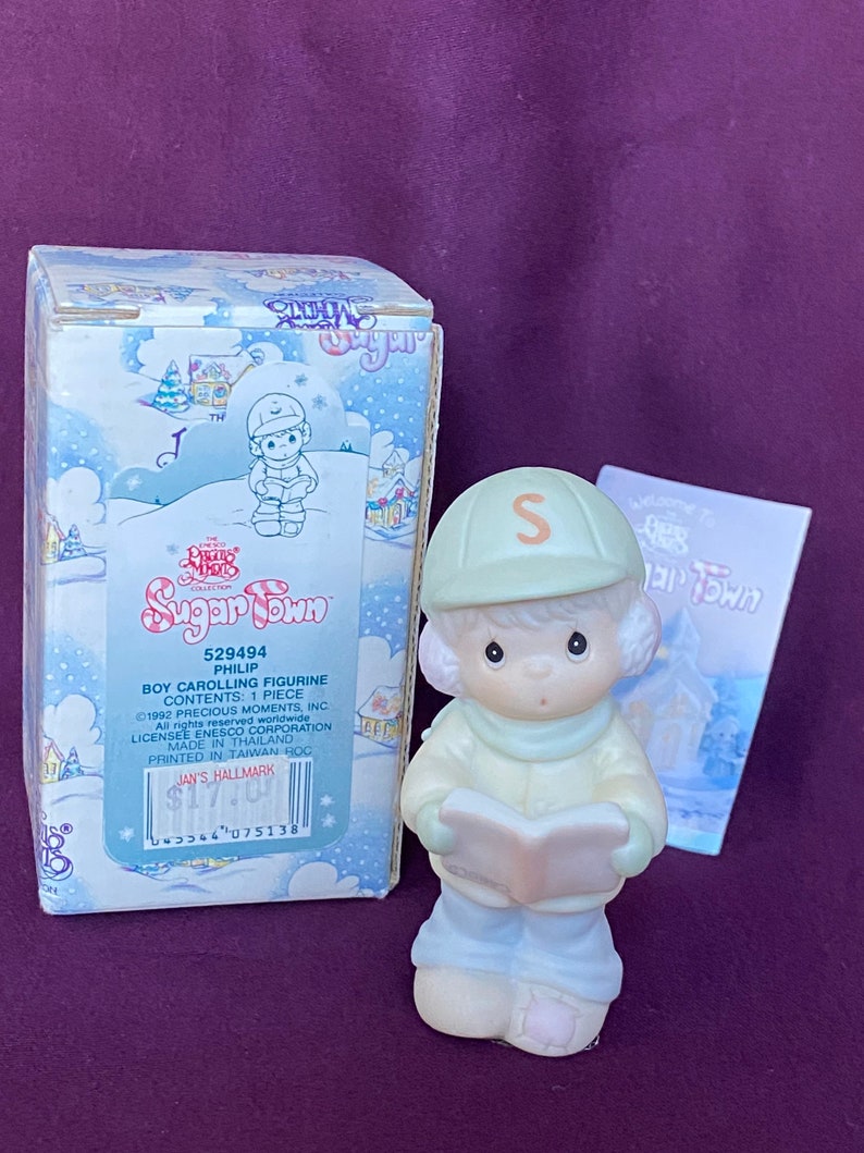 May include: A porcelain figurine of a boy wearing a green hat with the letter "S" on it, a blue jacket, and blue pants. He is holding a book and singing carols. The figurine is part of the Precious Moments Sugar Town collection.