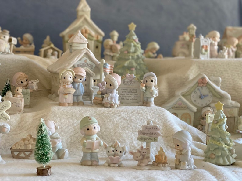 May include: A miniature Christmas village scene with a schoolhouse, a clock tower, a Christmas tree, and several figurines. The figurines are dressed in winter clothing and are holding gifts and other holiday items.