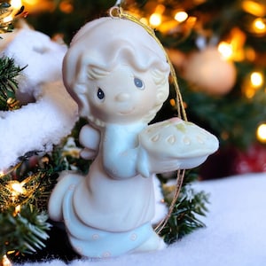 May include: A ceramic angel Christmas ornament, holding a pie. The angel is wearing a light blue dress and has a halo. The ornament is hanging in front of a Christmas tree with lights and snow.