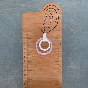 May include: A pair of earrings with a light purple and white circular design. The earrings are hanging from a wooden display stand with a ruler and the text "PANTHERA PRESS".