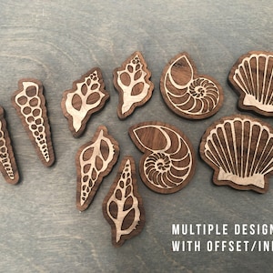Puede incluir: Un conjunto de nueve formas de conchas marinas de madera con intrincados diseños cortados con láser. Las conchas están hechas de madera de nogal y tienen un color marrón natural. Los diseños están desplazados e incrustados, creando un efecto 3D. "MULTIPLE DESIGNS WITH OFFSET/INLAY" está escrito en blanco en la parte inferior de la imagen.