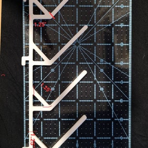 Może przedstawiać: Biały plastikowy szablon do pikowania z kątowymi ramionami, o wymiarach 3,18 cm i 6,99 cm. Szablon umieszczono na czarnej macie do cięcia z liniami siatki i pomiarami.