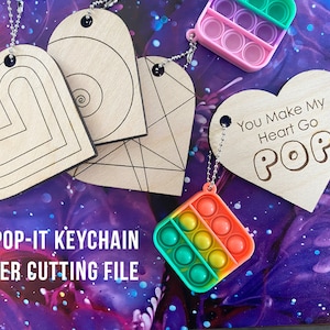 Puede incluir: Llavero de corazón de madera cortado con láser con un llavero pop-it adjunto. El corazón tiene el texto "You Make My Heart Go POP" grabado. El llavero pop-it es de colores arcoíris.
