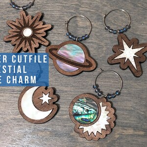 May include: Set of five laser cut wooden wine glass charms with celestial designs. The charms feature a sun, a planet, a star, a crescent moon, and a sun and moon combination. The charms are made of wood with a white inlay.