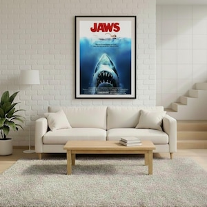 Puede incluir: Un p&oacute;ster enmarcado de la pel&iacute;cula "Jaws" cuelga sobre un sof&aacute; color crema. El p&oacute;ster presenta un tibur&oacute;n con la boca abierta, un nadador y el t&iacute;tulo "JAWS" en rojo. La habitaci&oacute;n tiene una paleta de colores neutros con una alfombra y una mesa de centro de madera.