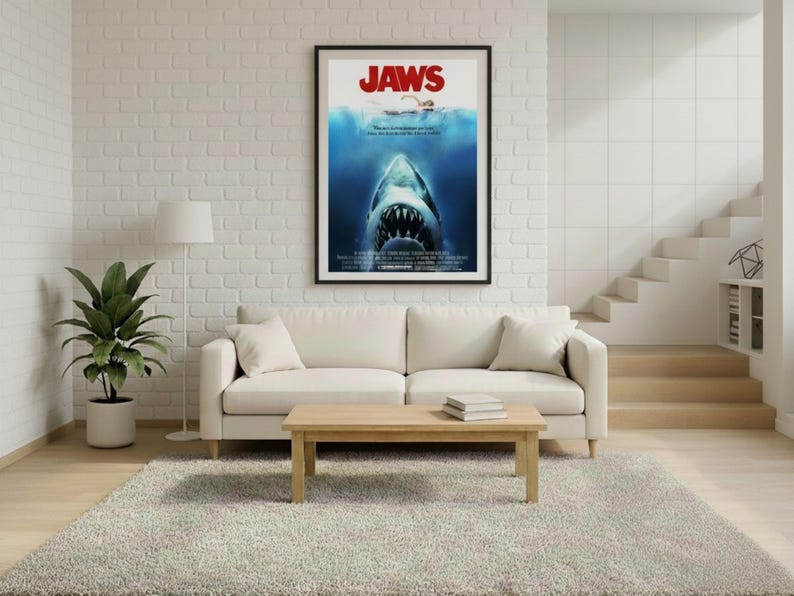 Puede incluir: Un p&oacute;ster enmarcado de la pel&iacute;cula "Jaws" cuelga sobre un sof&aacute; color crema. El p&oacute;ster presenta un tibur&oacute;n con la boca abierta, un nadador y el t&iacute;tulo "JAWS" en rojo. La habitaci&oacute;n tiene una paleta de colores neutros con una alfombra y una mesa de centro de madera.