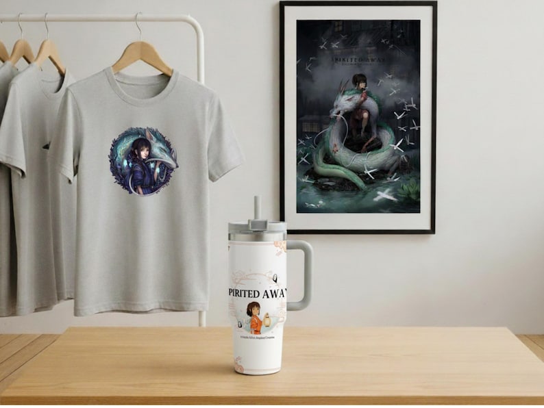 May include: A light gray t-shirt with a circular graphic featuring a character and a dragon. A white tumbler with a handle and the words "Spirited Away" is on a wooden surface. A framed print of a dragon and a person is on the wall.