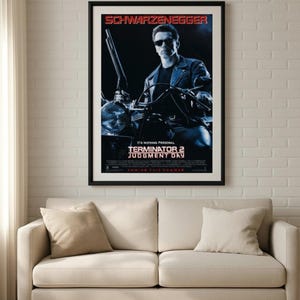 Puede incluir: P&oacute;ster enmarcado de la pel&iacute;cula "Terminator 2: El Juicio Final" con Arnold Schwarzenegger. El p&oacute;ster tiene un marco negro y est&aacute; colgado sobre un sof&aacute; beige. El t&iacute;tulo y el nombre del actor son en rojo y blanco.