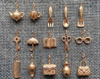 May include: A collection of fifteen gold-toned metal charms featuring various shapes including a teapot, teacup, fork, spoon, spatula, key, umbrella, open book, scissors, eyeglasses, comb, lightbulb, chest, vase, and a quilted handbag.