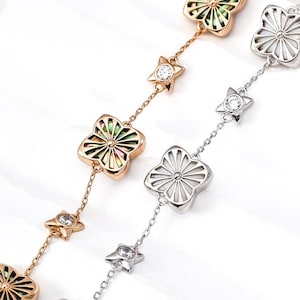 Puede incluir: Dos pulseras delicadas con un patrón repetitivo de dijes de flores de cuatro pétalos y pequeños acentos en forma de estrella. Una pulsera es de oro rosa con incrustaciones de concha iridiscente y la otra es de plata con incrustaciones blancas.