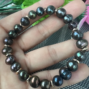 Może przedstawiać: Bransoletka z ciemnoszarych pereł ze złotymi przekładkami. Perły mają połysk i są nawleczone, tworząc okrągły kształt. Bransoletka jest trzymana w dłoni.