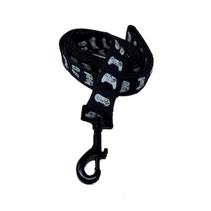 Puede incluir: Correa de perro negra con un patrón de gamepad blanco. La correa tiene un clip de metal negro.