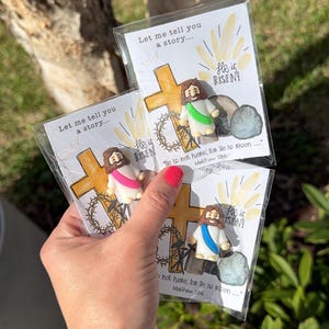 May include: Cards with a religious theme, featuring a cartoon depiction of Jesus, a cross, and the text "He is Risen!" The cards are packaged in clear plastic and held in a hand. The text "Let me tell you a story..." is also visible.