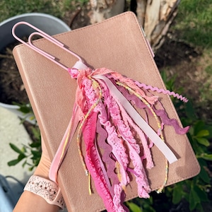 May include: A rose gold Bible with a pink paperclip bookmark and a tassel of pink, purple, and yellow ribbons and lace. The Bible is held in a person's hand.