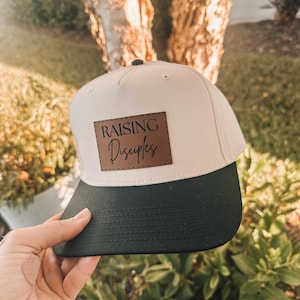May include: A beige and black baseball cap with the words "RAISING Disciples" on a brown leather patch. The cap has a curved brim and a button on top. The cap is being held up by a hand.
