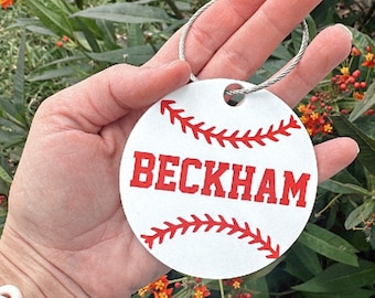 Puede incluir: Llavero redondo blanco con un diseño de béisbol rojo y el nombre "BECKHAM" en letras rojas.