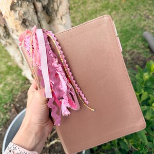 May include: A dusty rose-colored book with a matching bookmark and a decorative tassel of pink ribbons and lace. The book is held in a hand, with a pink floral watch band visible in the foreground.