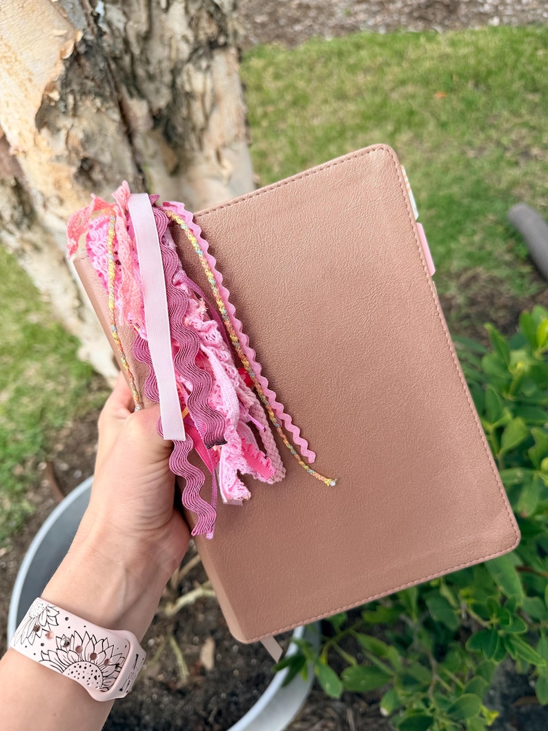 May include: A dusty rose-colored book with a matching bookmark and a decorative tassel of pink ribbons and lace. The book is held in a hand, with a pink floral watch band visible in the foreground.