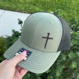May include: Olive green trucker hat with a black mesh back and a brown cross emblem. The hat has a curved brim and a tag that reads "RIC 1970".
