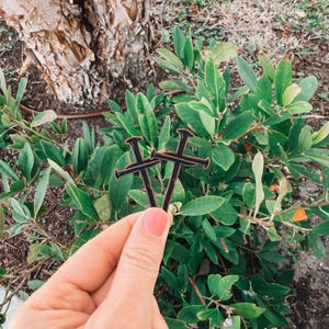 May include: Two black wooden cross-shaped ornaments with a brown border are held in front of green foliage. The ornaments are held by a person with pink nail polish. The background includes a tree trunk and more greenery.