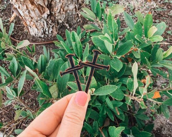 Puede incluir: Dos adornos de madera negros en forma de cruz con un borde marrón se sostienen frente a un follaje verde. Los adornos son sostenidos por una persona con esmalte de uñas rosa. El fondo incluye un tronco de árbol y más vegetación.