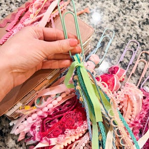 May include: A collection of colorful paperclip bookmarks with decorative ribbons and lace. The bookmarks feature various shades of pink, green, and blue ribbons, with pom-poms and lace accents. A brown leather journal is visible in the background.
