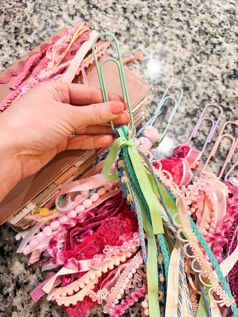 May include: A collection of colorful paperclip bookmarks with decorative ribbons and lace. The bookmarks feature various shades of pink, green, and blue ribbons, with pom-poms and lace accents. A brown leather journal is visible in the background.