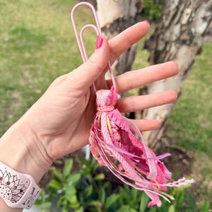 May include: A pink paperclip with a decorative tassel. The tassel is made of various shades of pink fabric strips and ribbons. The paperclip is held in a hand with pink nail polish, with a pink watch band with a sunflower design in the background.