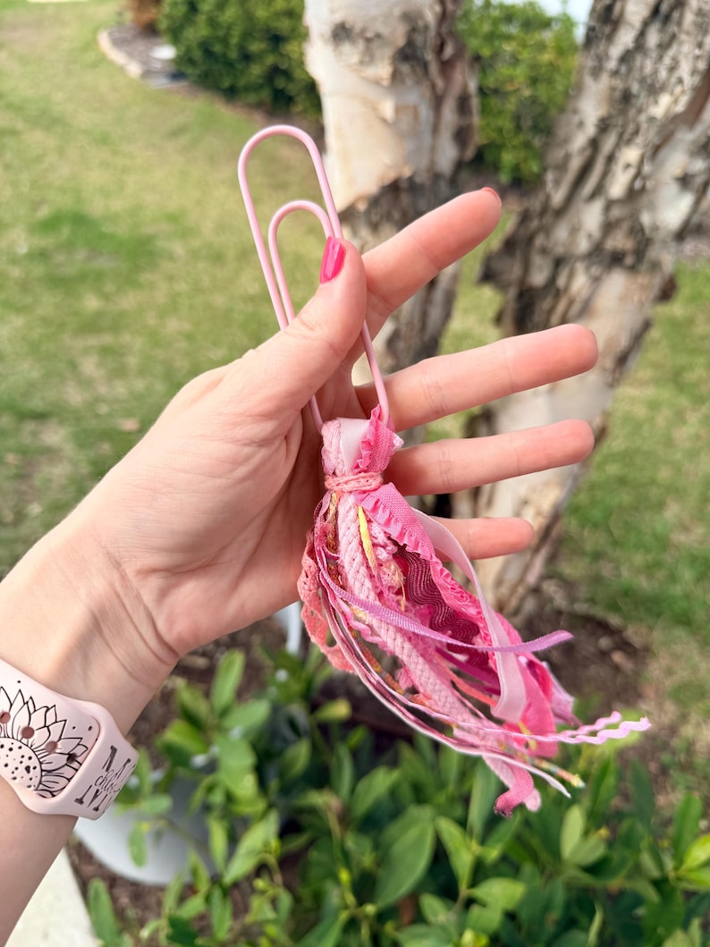 May include: A pink paperclip with a decorative tassel. The tassel is made of various shades of pink fabric strips and ribbons. The paperclip is held in a hand with pink nail polish, with a pink watch band with a sunflower design in the background.