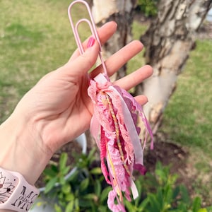 May include: A pink paperclip holds a tassel of pink and white ribbons, lace, and gold thread. The tassel is a mix of textures and shades of pink, creating a decorative accent. The paperclip is held in a hand.