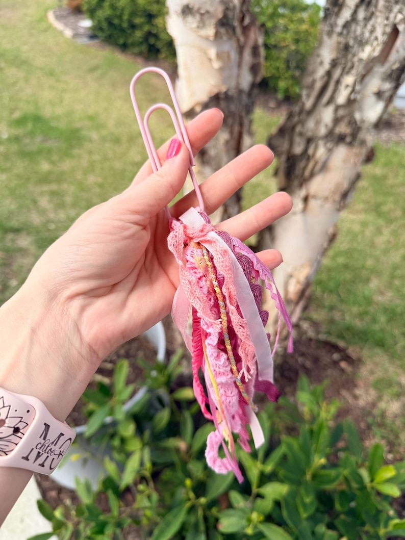 May include: A pink paperclip holds a tassel of pink and white ribbons, lace, and gold thread. The tassel is a mix of textures and shades of pink, creating a decorative accent. The paperclip is held in a hand.