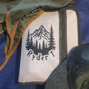 May include: A light-colored zippered case with a mountain and pine tree graphic and the name "Ryder T." printed in black. A green and brown fabric is visible in the background, along with a black cap.
