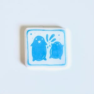 May include: A square, white ceramic tile with a hand-painted design. Two blue, cartoon-like figures are depicted, one larger than the other, with a splash of water or a similar element between them. The figures have simple features and are set against a light blue background.